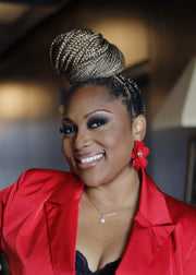 Woman wearing a red jacket with braided hair and bold, floral earrings, smiling.