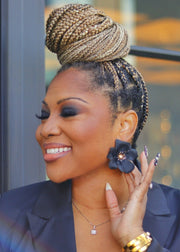 Woman with braided hair wearing a black power suit and statement flower earrings, standing in front of a blurred background.
