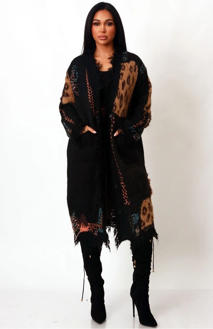 Woman wearing a black leopard distressed longline cardigan with pockets, styled as a coat over black outfit and knee-high boots.